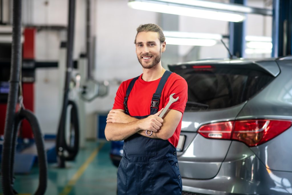 An automotive mechanic smiling and holding a wrench in a garage.